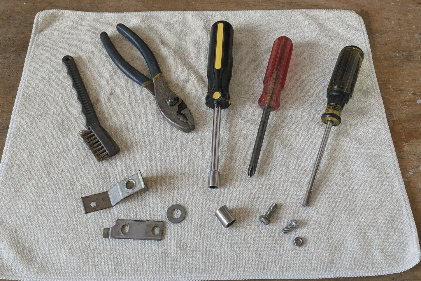 Wire brush, pliers, nut driver and screwdrivers laid on a towel with two steel brackets, a washer and loose screws